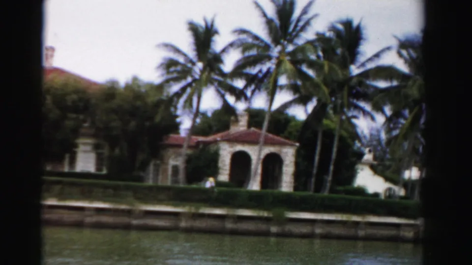 1955 Dade County waterfront frame showing palms, an arched porch, hedges, canal water, and a small distant person near private property.