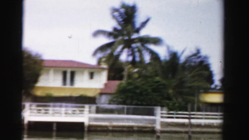 1955 Dade County 8mm frame showing a white waterfront house, dock railing, palms, dense shrubs, and the edge of the film frame.