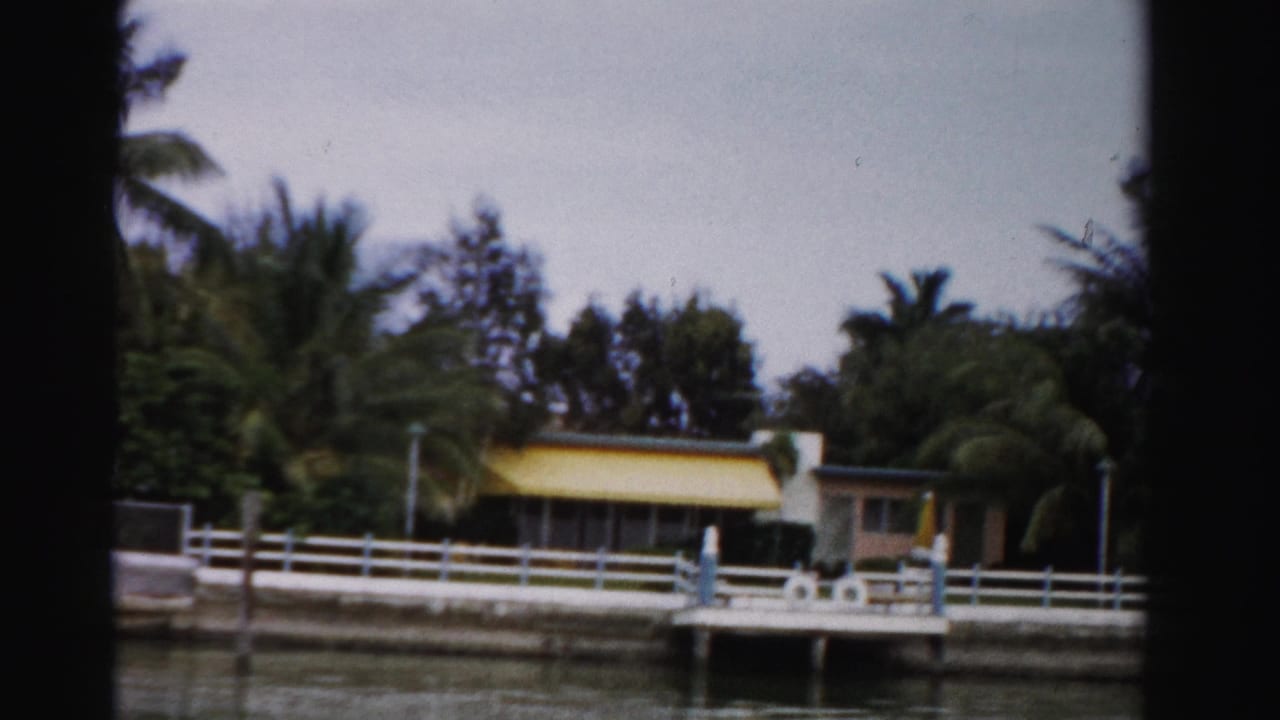 Dade County canal-side residence with yellow awning, white railing, dock structure, and tropical palms in 1955 footage.