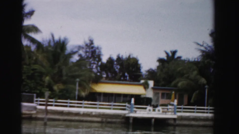 Dade County canal-side residence with yellow awning, white railing, dock structure, and tropical palms in 1955 footage.