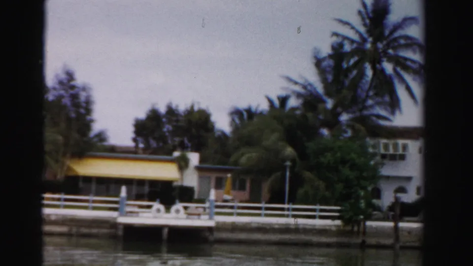 1955 Dade County waterfront property seen from the water, with white fencing, a dock, palms, and a low building with yellow awning.