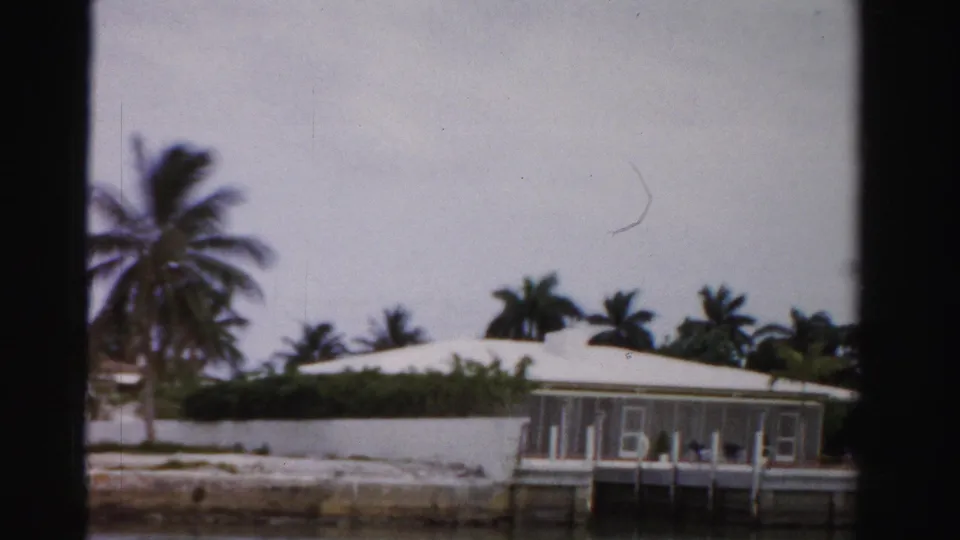 Dade County waterfront home with a light roof, dock, seawall, and palms, viewed from across canal water in 1955 8mm footage.