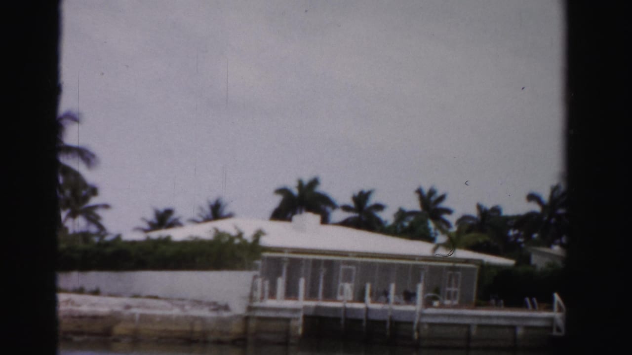 1955 Dade County, Florida view from a boat of a white waterfront house with screened porch, palms, seawall, and calm water.