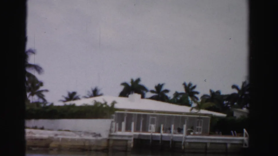 1955 Dade County, Florida view from a boat of a white waterfront house with screened porch, palms, seawall, and calm water.