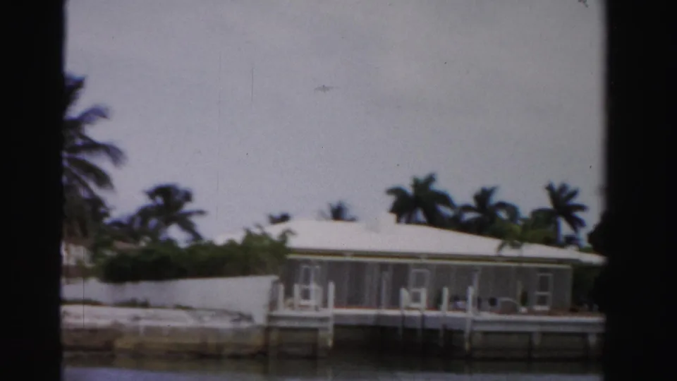 Boat-level 1955 Dade County view of a white canal-side house with screened porch, dock pilings, palms, and a pale cloudy sky.