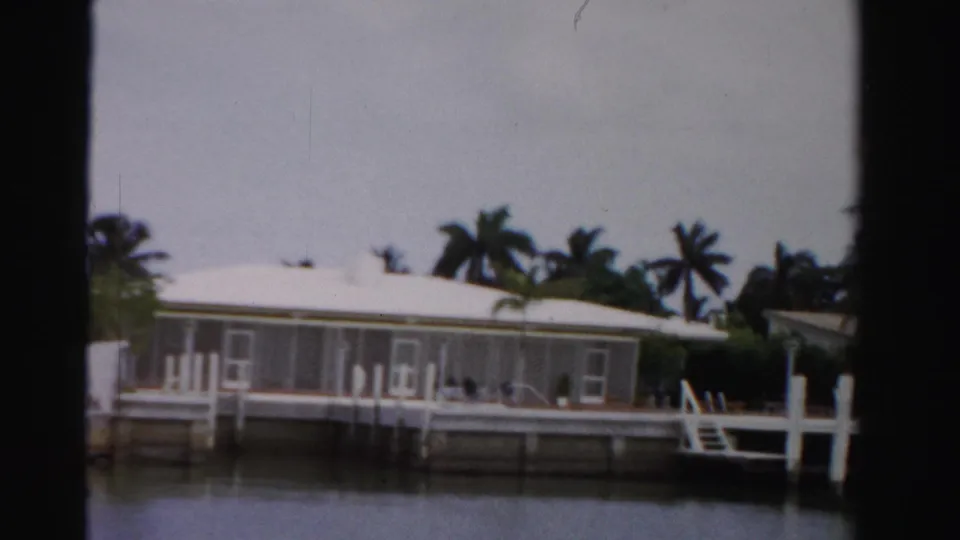 1955 Dade County waterfront residence seen from a moving boat, with screened windows, dock structure, fence posts, palms, and still water.