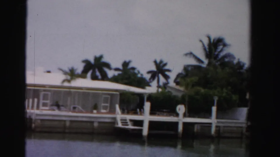 Final 1955 Dade County boat-view frame of a white waterfront house partly obscured by dock posts, fence details, palms, greenery, and water.