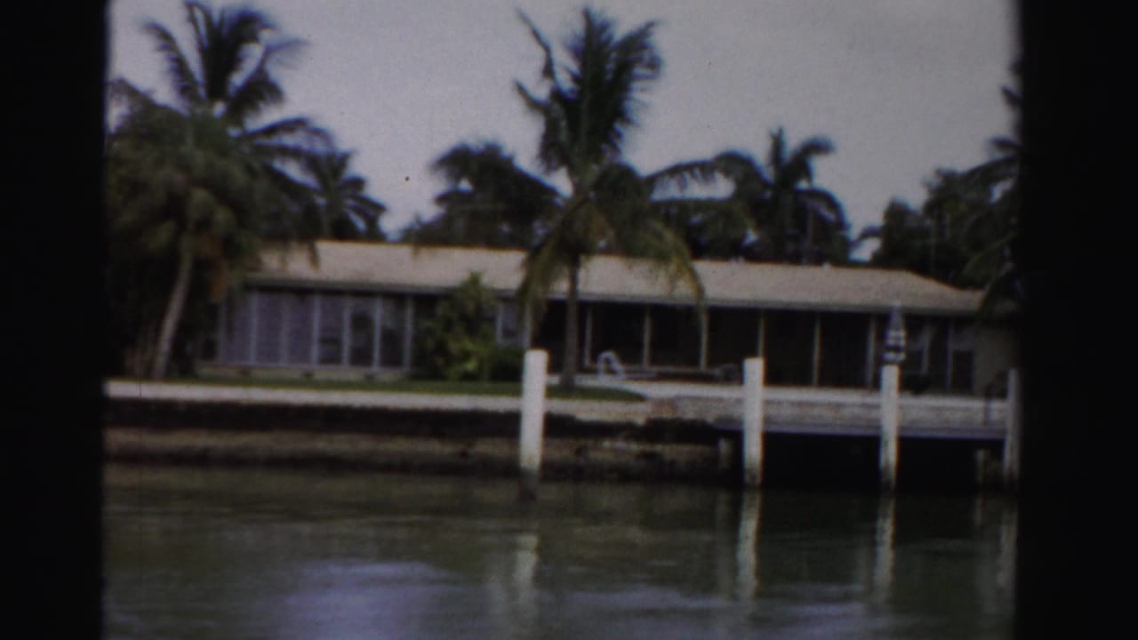 1955 Dade County 8mm frame showing a low canal-front residence with palms, white pilings, seawall, and water reflections.
