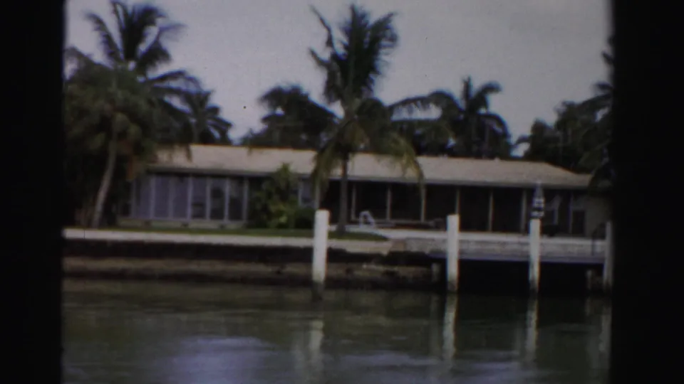 1955 Dade County 8mm frame showing a low canal-front residence with palms, white pilings, seawall, and water reflections.
