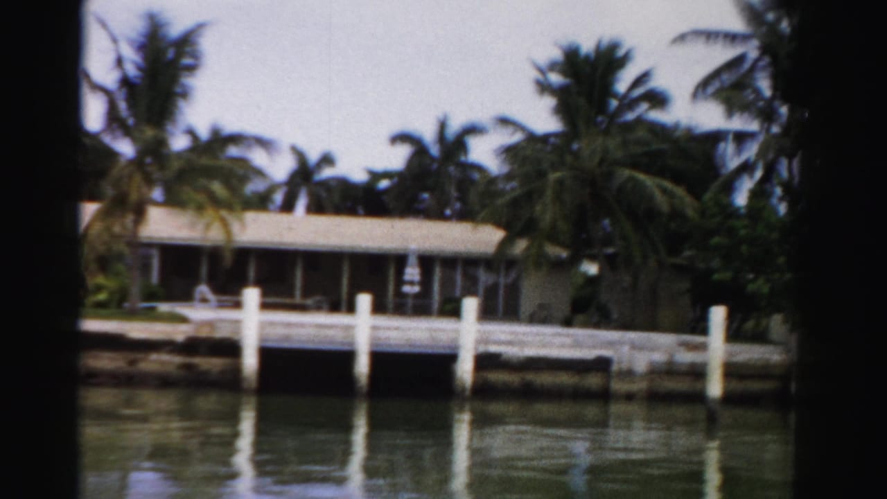 1955 Dade County home-movie frame of a canal-front house, palm landscaping, white dock pilings, and soft water reflections.