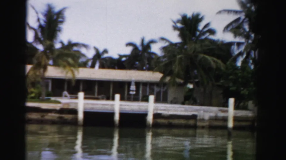 1955 Dade County home-movie frame of a canal-front house, palm landscaping, white dock pilings, and soft water reflections.