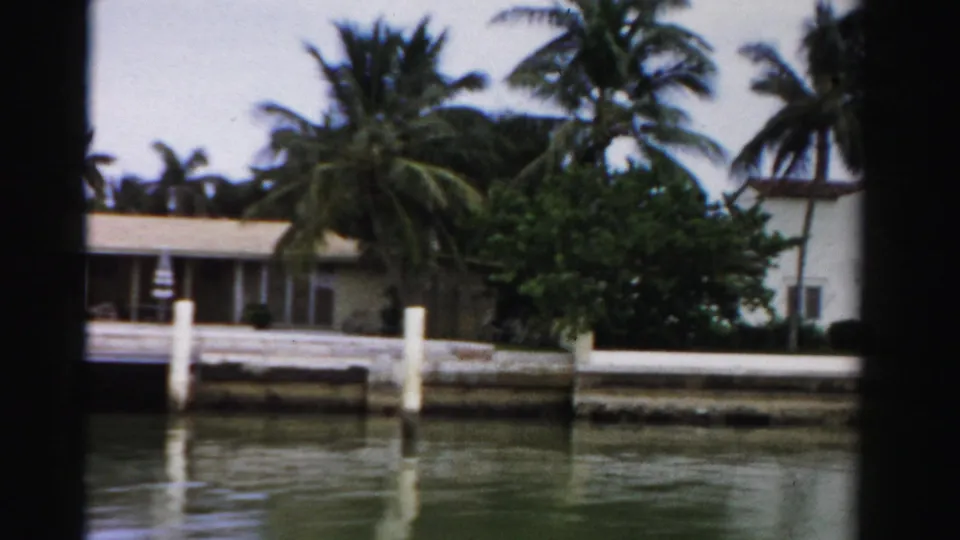 Dade County 1955 boat-view frame showing a palm-shaded waterfront house, white dock posts, seawall, and green canal water.