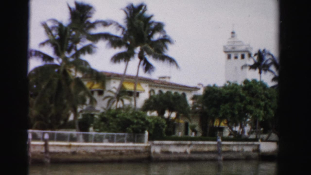 1955 Dade County waterfront view of a large building with yellow awnings, palms, a tower, seawall, and calm water in the foreground.