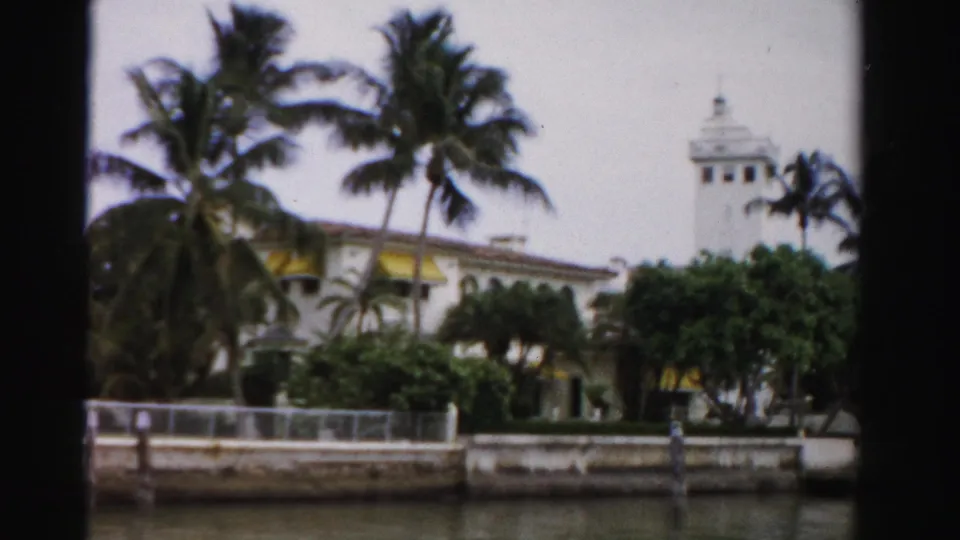 1955 Dade County waterfront view of a large building with yellow awnings, palms, a tower, seawall, and calm water in the foreground.