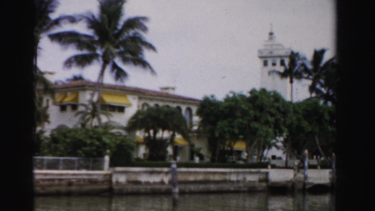 Palm-lined waterfront building with yellow awnings, arched windows, seawall, and tower.