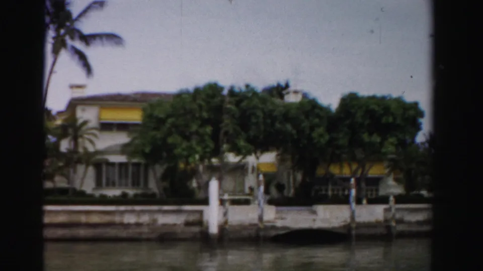 1955 Dade County waterfront building partly screened by trees, with yellow awnings, white posts, a seawall, and water in the foreground.