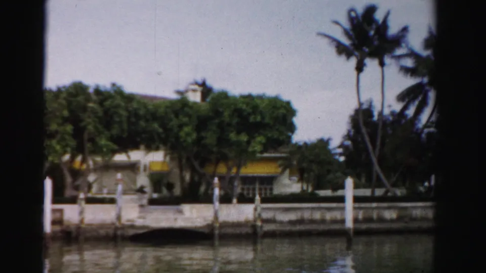 1955 Dade County waterfront scene with a tree-covered building, yellow awnings, palms, white posts, seawall, and calm water reflections.
