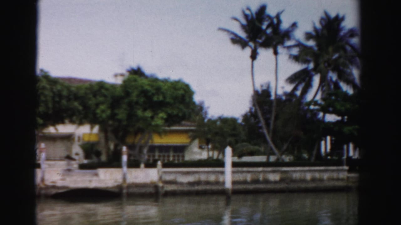 1955 Dade County canal view of a waterfront residence with palms, a seawall, white dock posts, and a yellow awning.