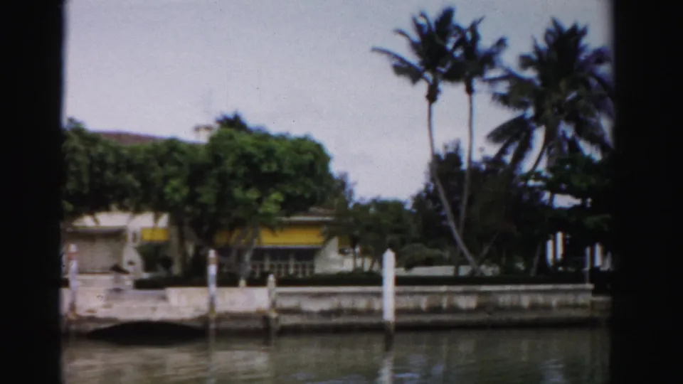 1955 Dade County canal view of a waterfront residence with palms, a seawall, white dock posts, and a yellow awning.