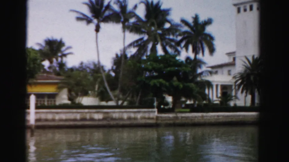1955 Dade County canal-side residence framed by palms, tropical shrubs, a seawall, white posts, and muted 8mm color.