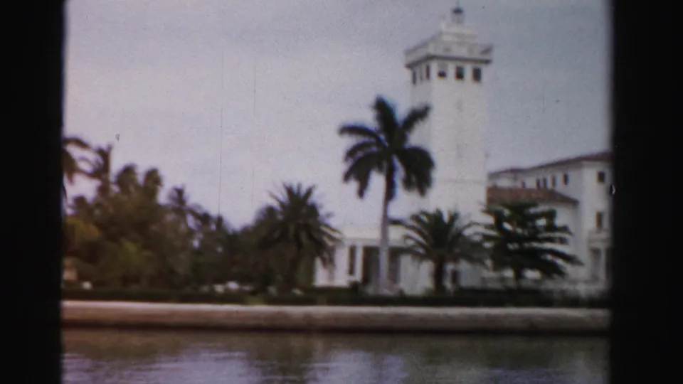 1955 Dade County view of a prominent white waterfront building with a tower, palms, landscaping, and canal water in the foreground.