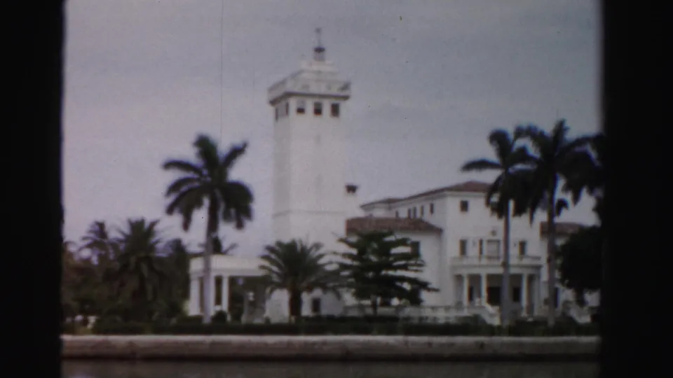 1955 Dade County waterfront view of a white estate with a square tower, balcony, palm trees, and low seawall.