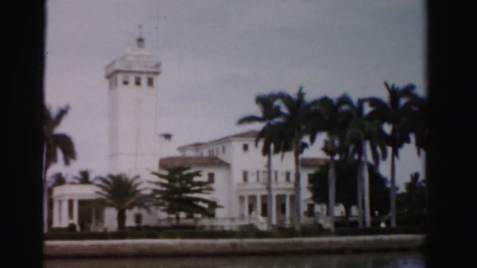 1955 Dade County 8mm view of the waterfront mansion, with tower and entrance area partly hidden by palms and dark frame edges.