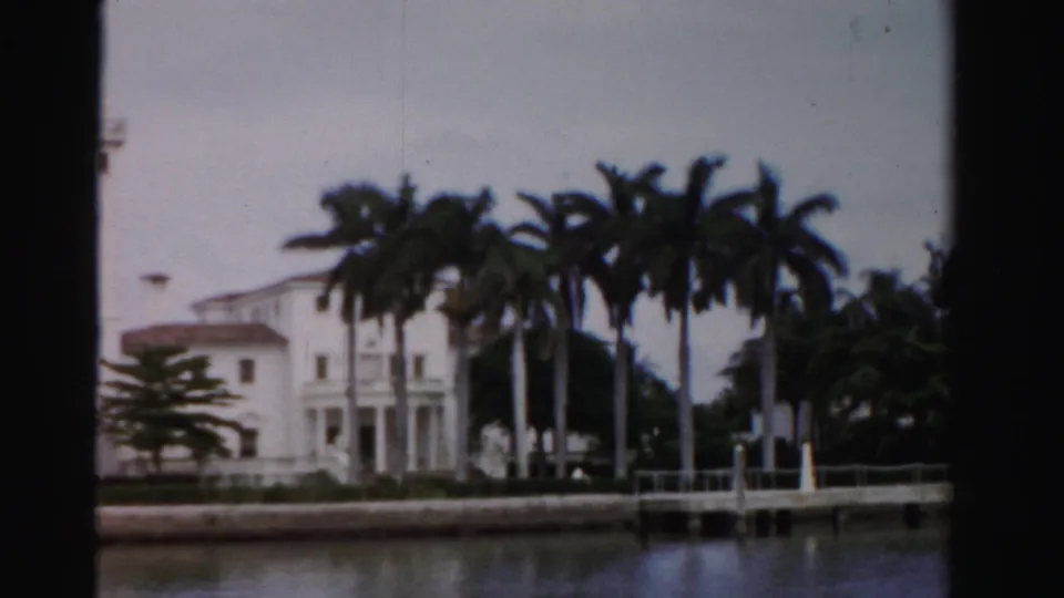 1955 Dade County 8mm frame showing a white waterfront residence, palms, dock posts, and calm canal water.