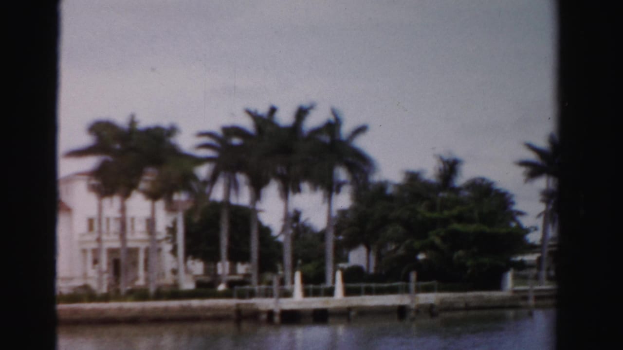 1955 Dade County home-movie frame of a white waterfront house partly screened by palms beside a small dock.