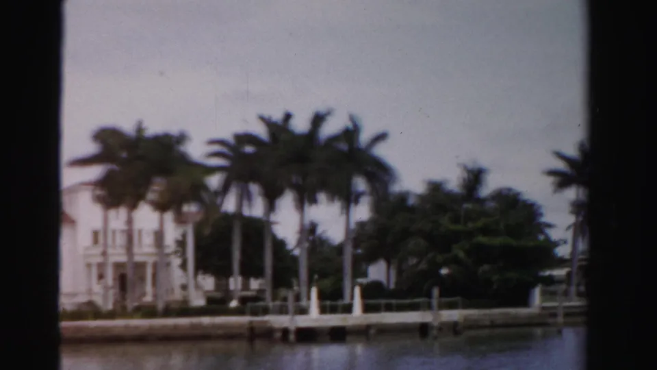 1955 Dade County home-movie frame of a white waterfront house partly screened by palms beside a small dock.