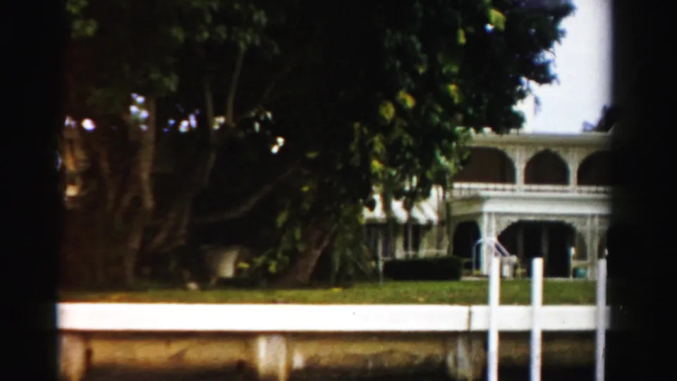1955 Dade County 8mm frame showing a large white house with an arched porch, lawn, trees, and canal-side posts.