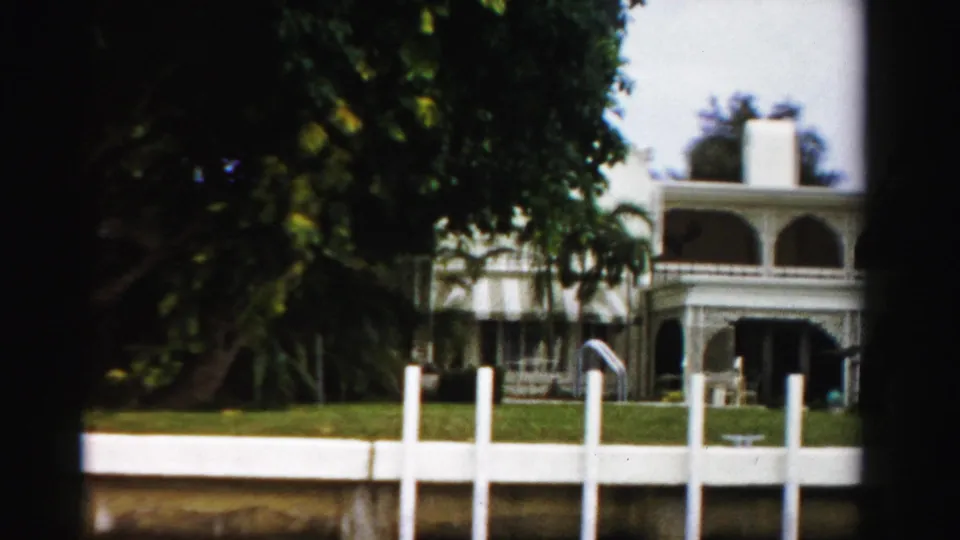 1955 Dade County home-movie frame of a white arched waterfront house with palms, lawn, dock posts, and dense foliage.