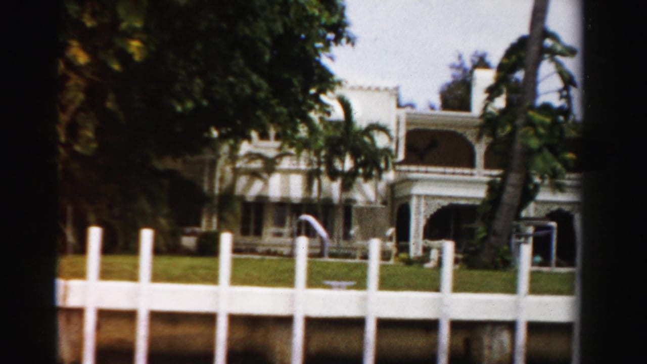 1955 Dade County 8mm frame showing a white mansion with arched porches, palm trees, lawn, and waterfront-style fencing.