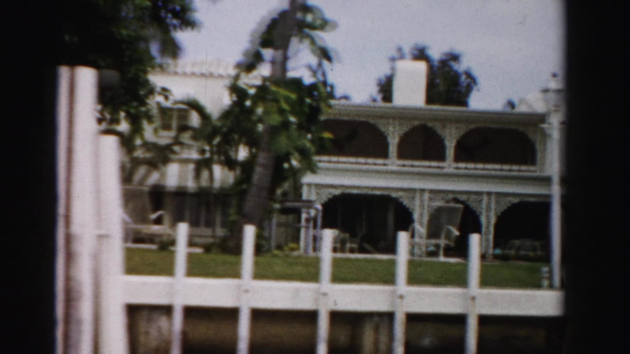 White fence and palm trees in front of a large arched mansion.