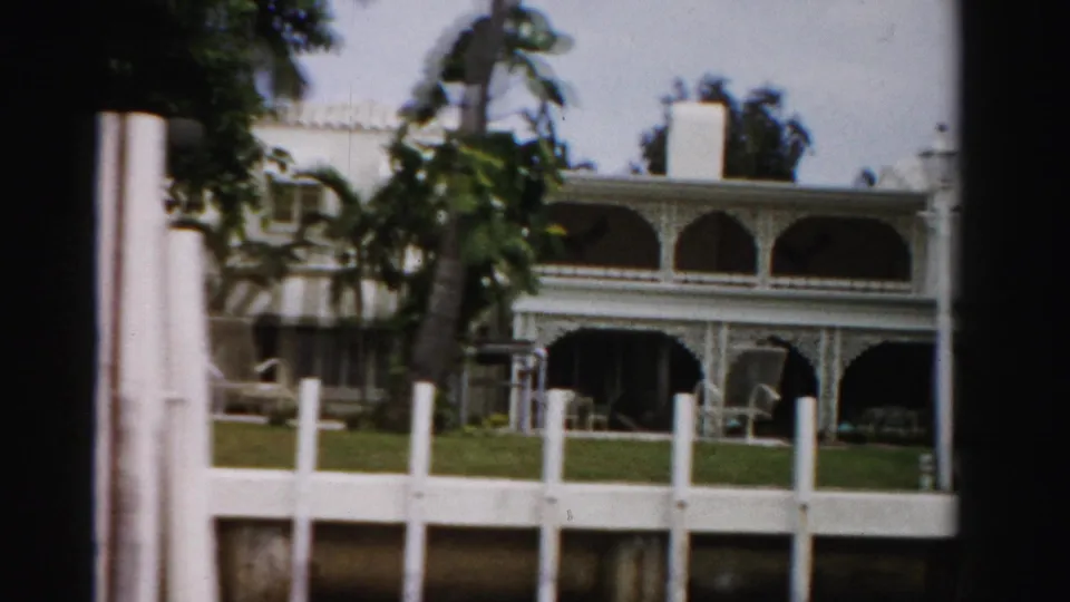 1955 Dade County frame with a large white arched mansion partly screened by palm trees and a white fence in the foreground.