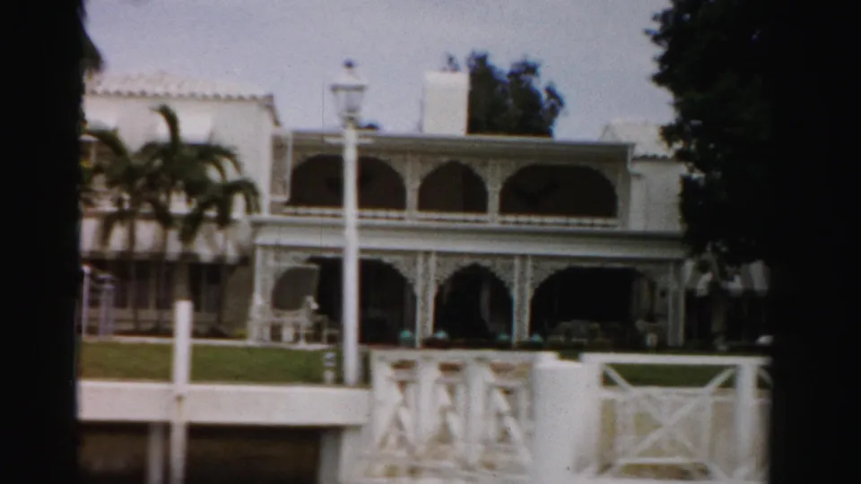 1955 Dade County 8mm view of the mansion facade, showing arched balconies, a lawn, white fence sections, and a lamppost.
