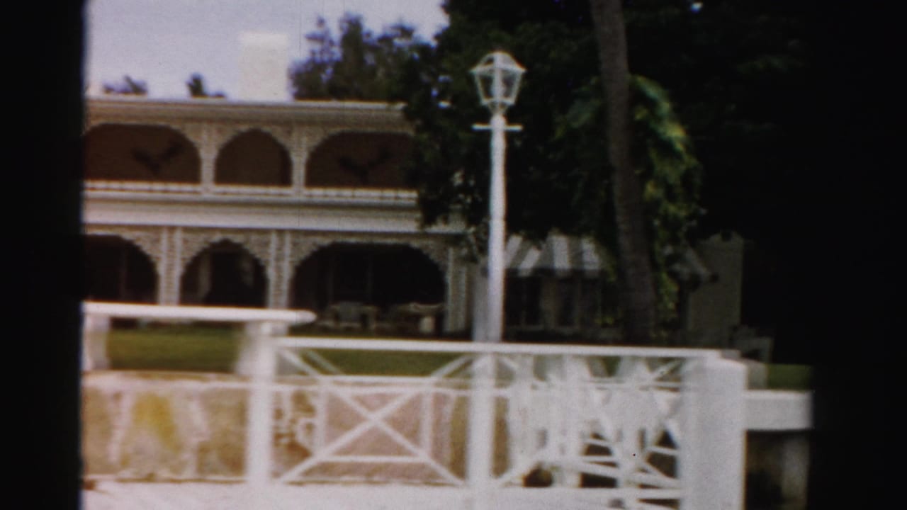 1955 Dade County 8mm frame showing a white oceanfront estate with arched veranda, palms, dock railing, lamppost, and seawall.