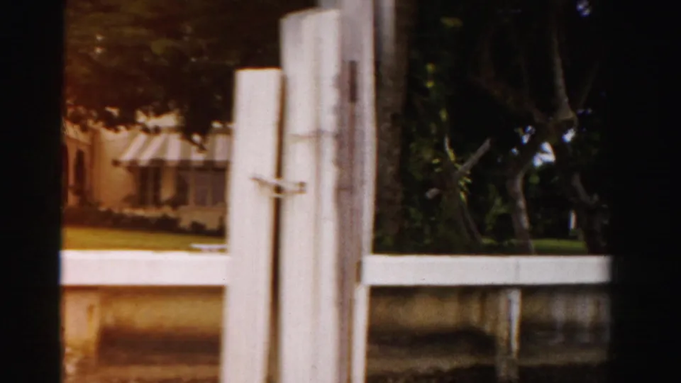 1955 Dade County 8mm frame from a moving viewpoint, showing a waterfront building, striped awning, white post, trees, dock edge, and seawall.