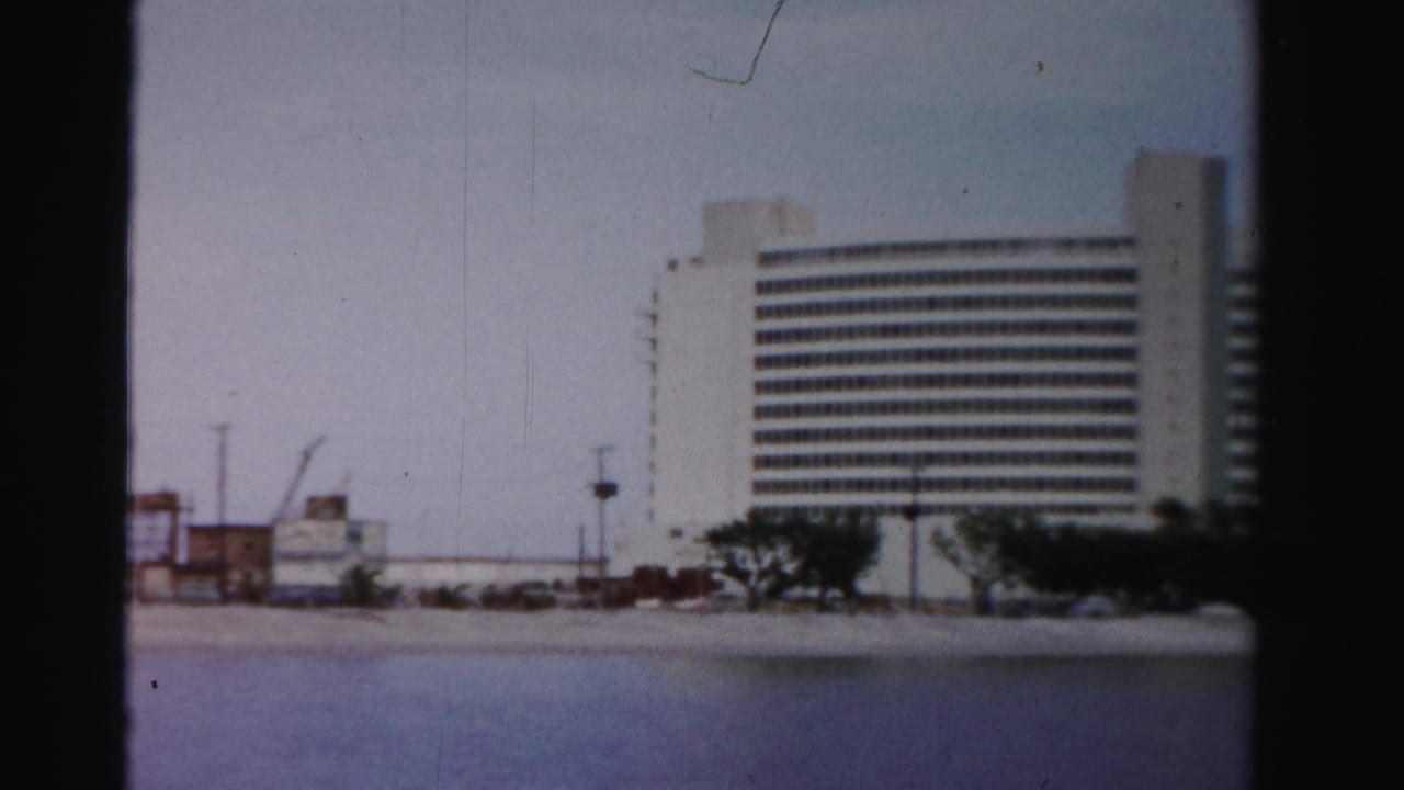 A white multi-story waterfront hotel or apartment building appears along the Dade County shoreline in soft 8mm color.