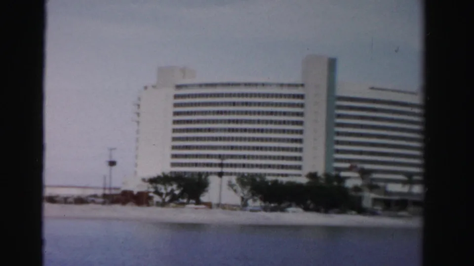 The waterfront building fills more of the frame, showing its curved facade, rows of windows, palms, and beach edge.