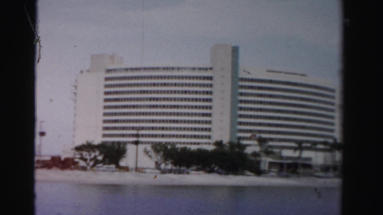 1955 Dade County home-movie frame showing a white modernist waterfront building with palms, shoreline, and cars in front.