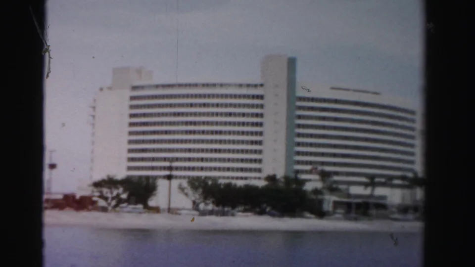 1955 Dade County home-movie frame showing a white modernist waterfront building with palms, shoreline, and cars in front.