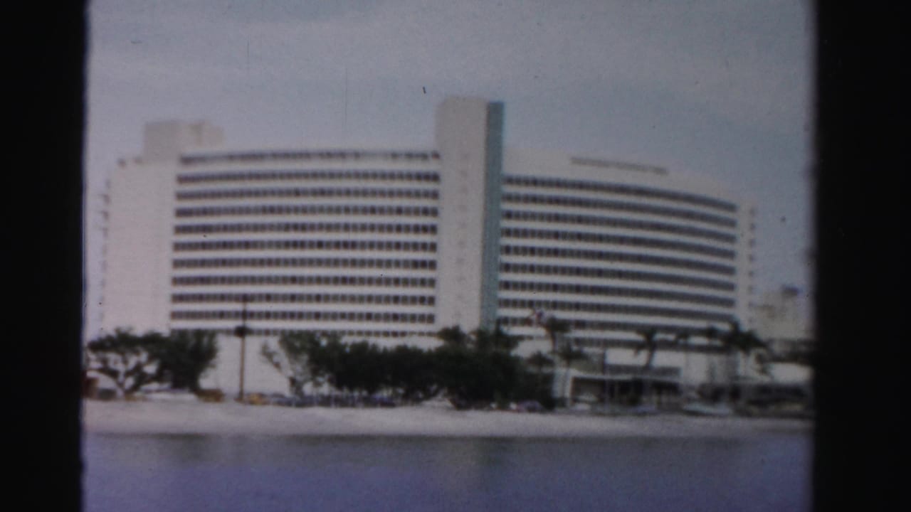1955 Dade County frame of a large white waterfront building with a curved facade, window grid, palms, and beachside road.