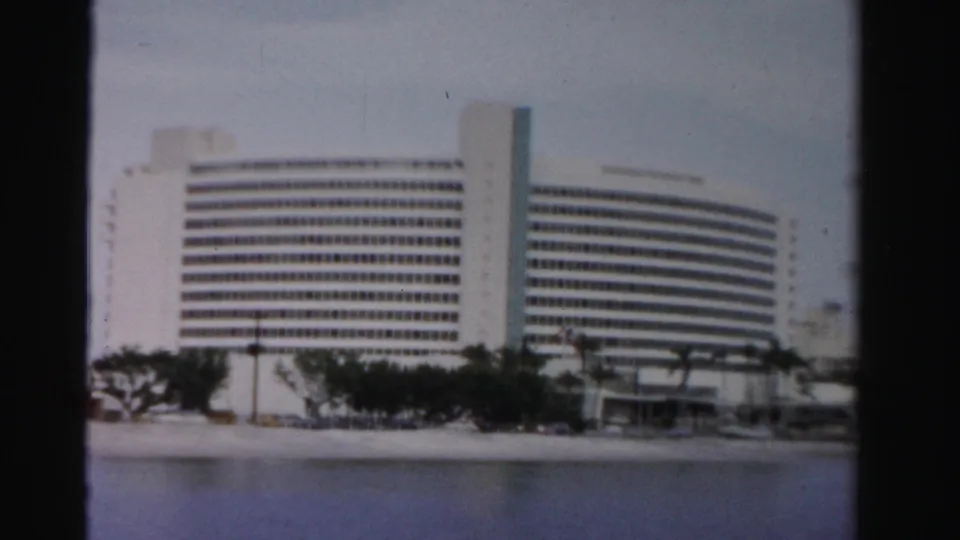 1955 Dade County frame of a large white waterfront building with a curved facade, window grid, palms, and beachside road.
