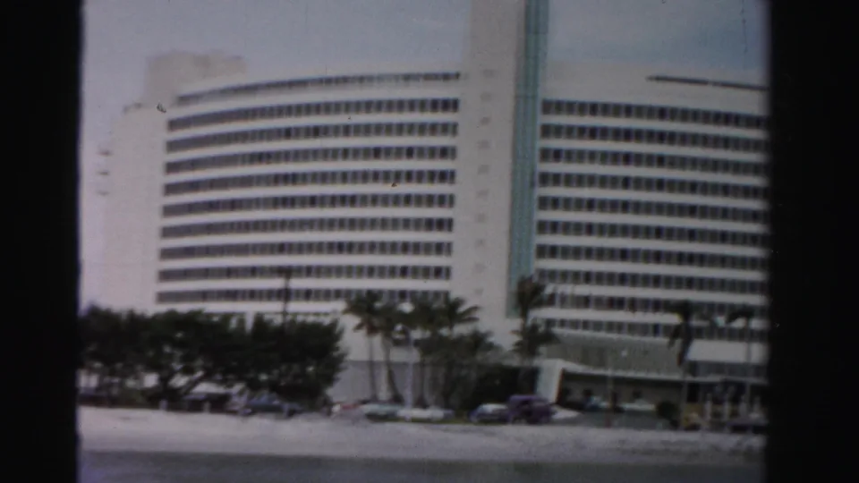 Closer 1955 Dade County view of the modernist waterfront building, showing its window grid, palms, parked cars, and entry area.