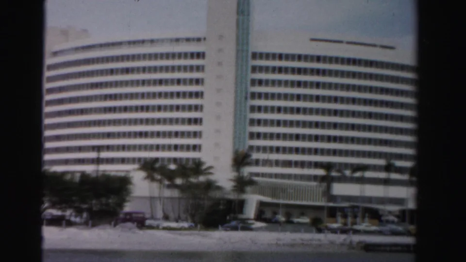 1955 Dade County home-movie frame showing a closer view of the white waterfront building, palms, cars, and dark 8mm frame borders.
