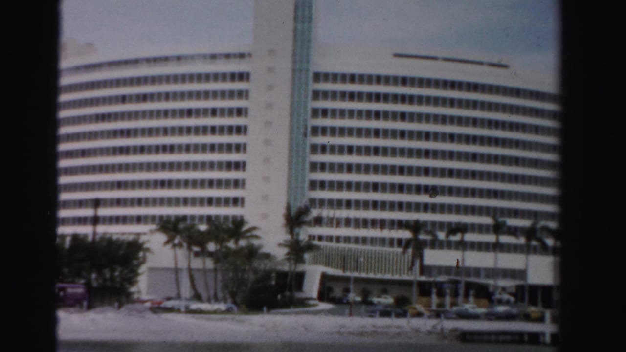 1955 Dade County home-movie view of a large white windowed high-rise with palms, cars, and a low foreground road or waterfront edge.