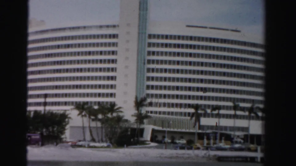 1955 Dade County home-movie view of a large white windowed high-rise with palms, cars, and a low foreground road or waterfront edge.