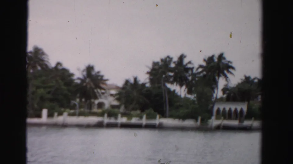 1955 Florida home-movie frame of waterfront residences, palms, a seawall, and arched dock structure across calm water.