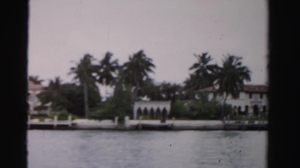 1955 Dade County 8mm view of a white Mediterranean-style waterfront house behind palm trees and dock posts.